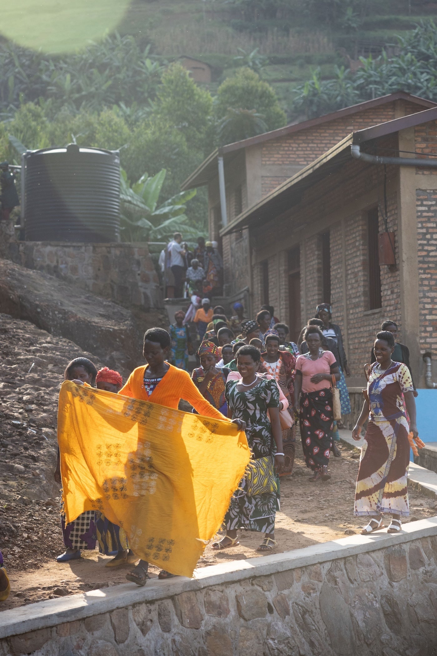 Group of people, including children, standing on a dirt path near a building with a water tank in the background.